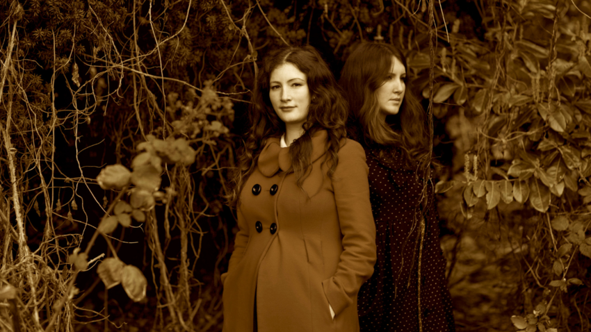 two women stand close to one another surrounded by branches and leafs