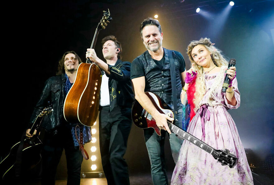 Four people standing on a stage, three holding gutairs and one holding a microphone