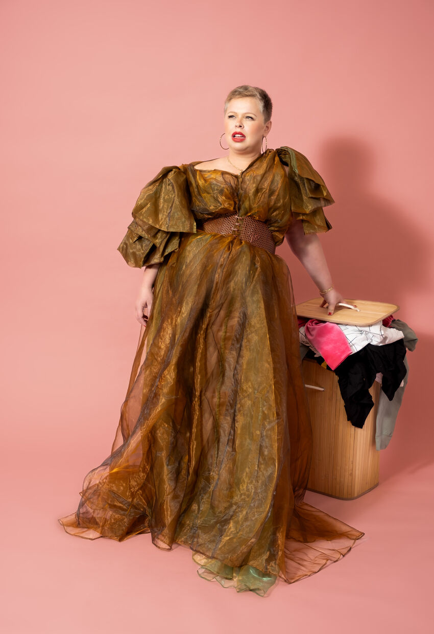A white woman with short blonde hair wearing a brown/gold pouffy dress, learning against an overflowing washing basket