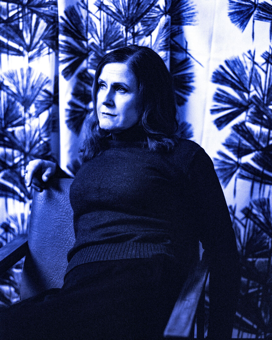 Photo of Alison Moyet with a blue tint filter sitting on a chair looking away from the camera, floral curtains in the background