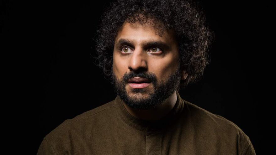 Nish Kumar looking dramatically away from camera in front of a dark background. He's a man of Indian descent in his late 30s wearing a simple brown shirt