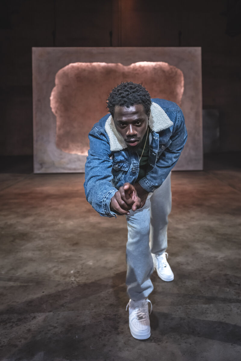 An actor wearing a light blue denim jacket crouches forward with his index finger extended as thought about to touch something.