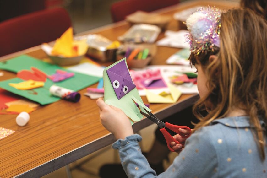 A young girl at a craft table cutting paper. She's wearing a sparkly headband