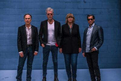 Four people standing in front of blue brick wall. They are wearing black blazers, jeans and all have their arms by their sides