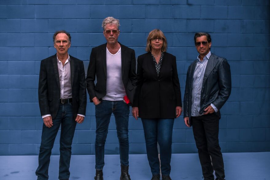 Four people standing in front of blue brick wall. They are wearing black blazers, jeans and all have their arms by their sides