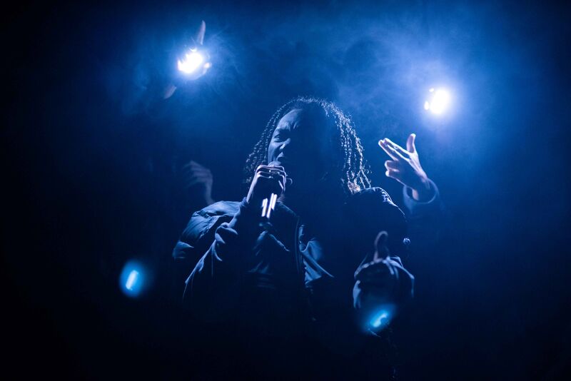 A man with shoulder length hair is lit from behind by bluie lights. He is shouting into a microphone making gun signs with his hands.