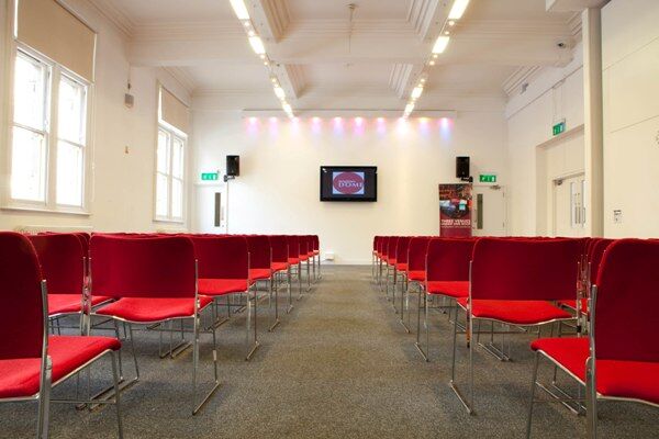 A white walled room with rows of red chairs layed out and a screen on the back wall.