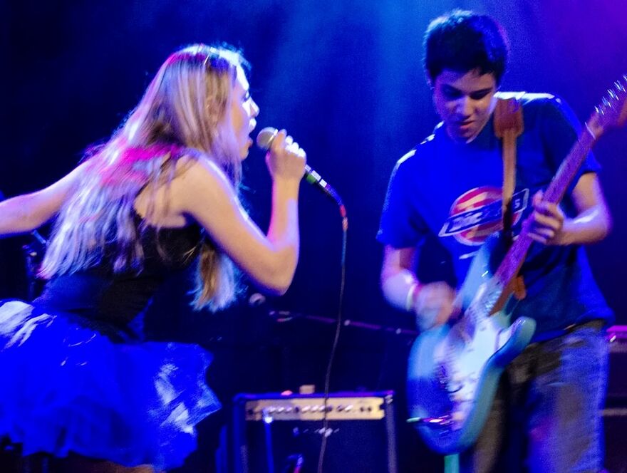 Two people on stage. One is singing into a microphone and the other is playing guitar