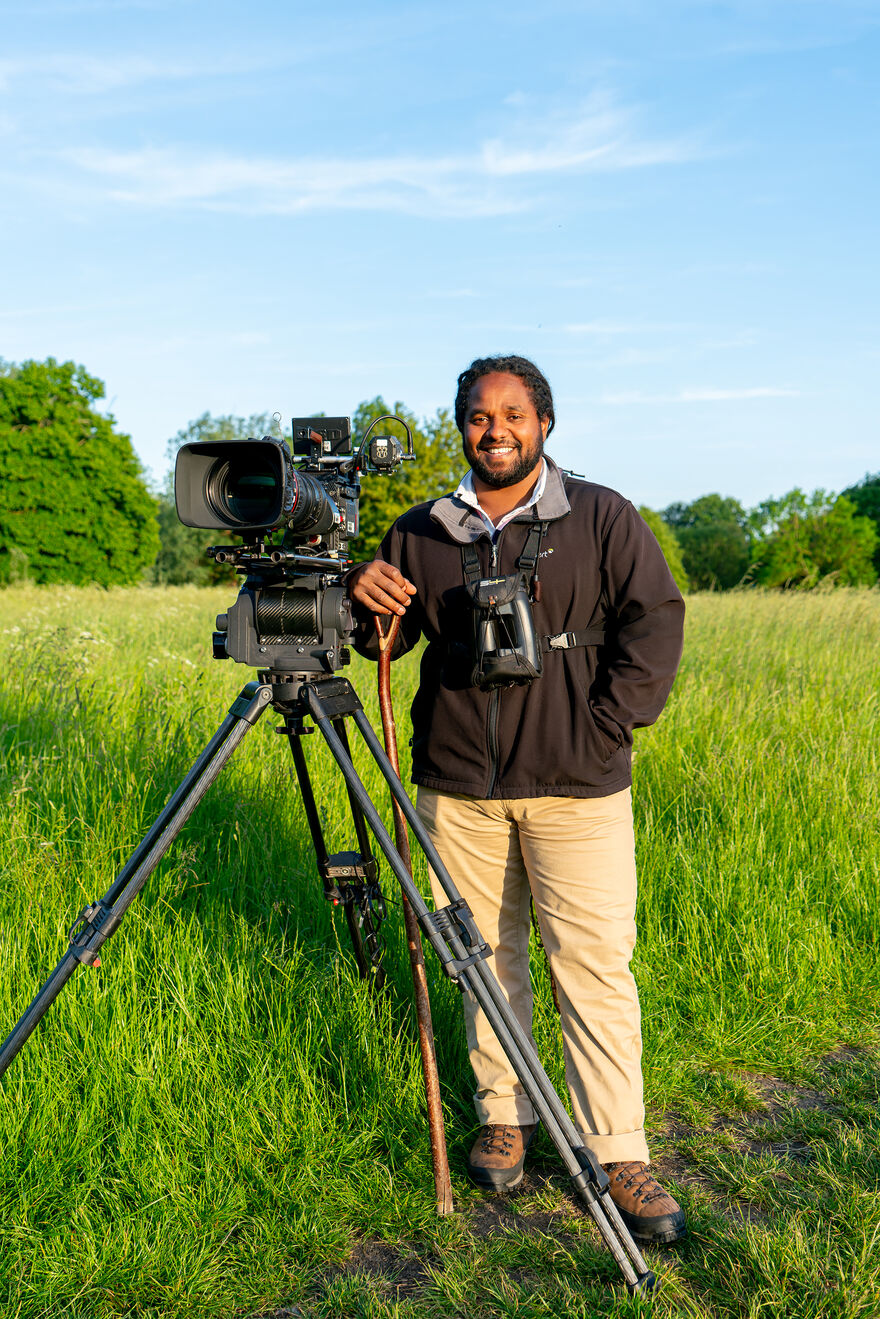 Hamza Yassin standing in a field with a camera propped on a tripod. He is smiling