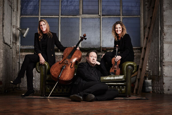 Two women and a mn dressed in smart black concert clothes posed around a green leather sofa. The women are holding a cello and a violin.