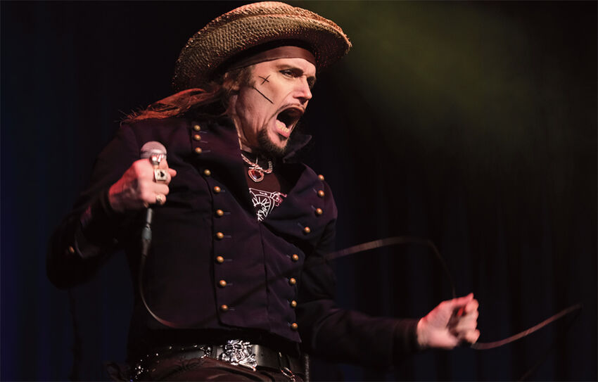 A white man dressed in a black shirt with two lines of gold buttons on it, plus a pirate-style hat. He is holding a microphone in his left hand and his mouth is wide open, as if he's singing.