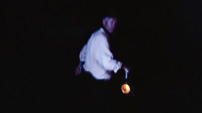 A white man in a white shirt, looks back over his shoulder whilst carrying an old railway torch light in his right hand