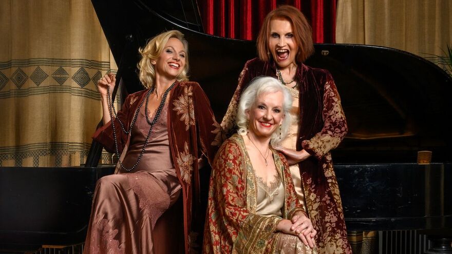Three women wearing brown and gold dresses and pearls in front of a grand piano. They are all smiling 