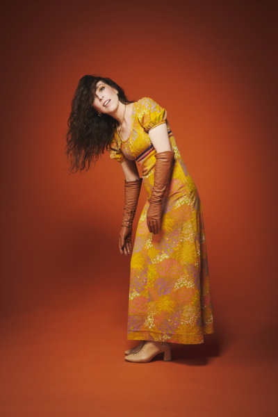 Bridget Christie in a yellow top standing over a brown background 