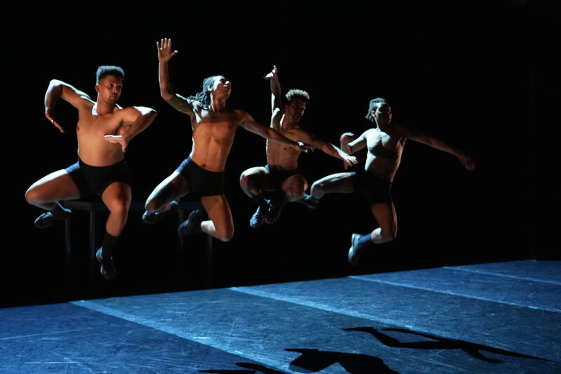 Four black male dancers jump on stage lit from the side by a single warm light which outlines their bodies