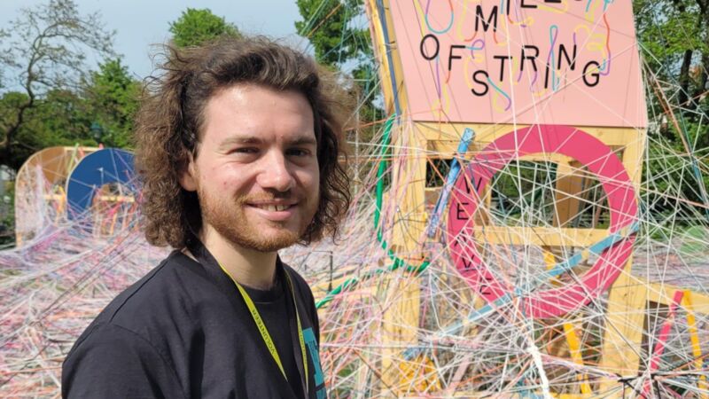 A white man stands outside by an interactive art installation. A sign reads 100 Miles of String, and there is lots of string crossing and weaving behind him