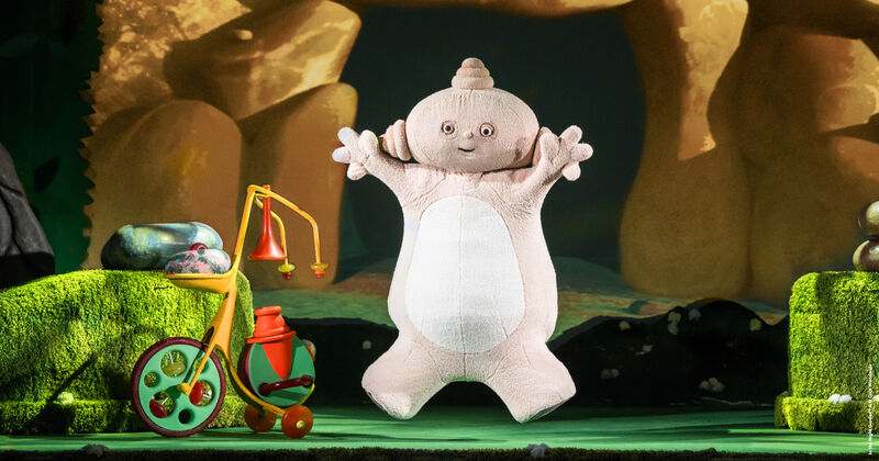 A beige costumed figure is mid-air in a star jump with a multi-coloured tricycle set beside it