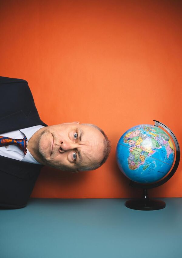 Jack Dee leaning into camera sideways, wearing a smart suit and a stern expression. Above his head is a globe