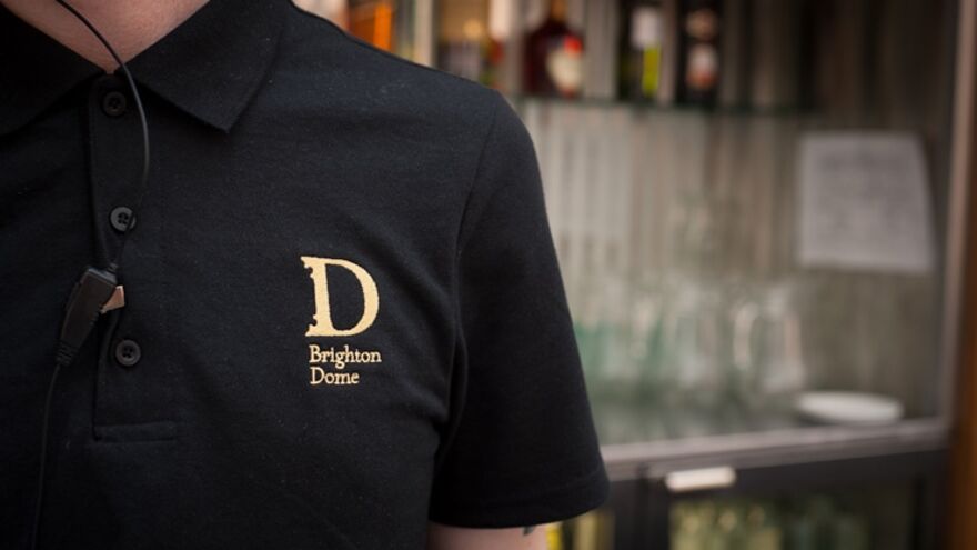 A person wearing a black polo shirt with a gold Brighton Dome logo on it