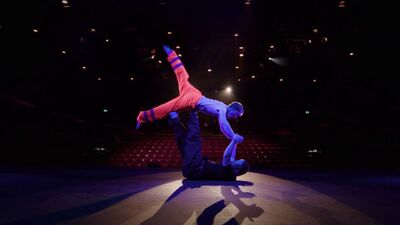 Two dancers are in a spotlight on a stage. In the background you can see the empty theatre auditorium. One dancer is in all black clothing, and the other has hi-vis orange trousers with reflective stripes