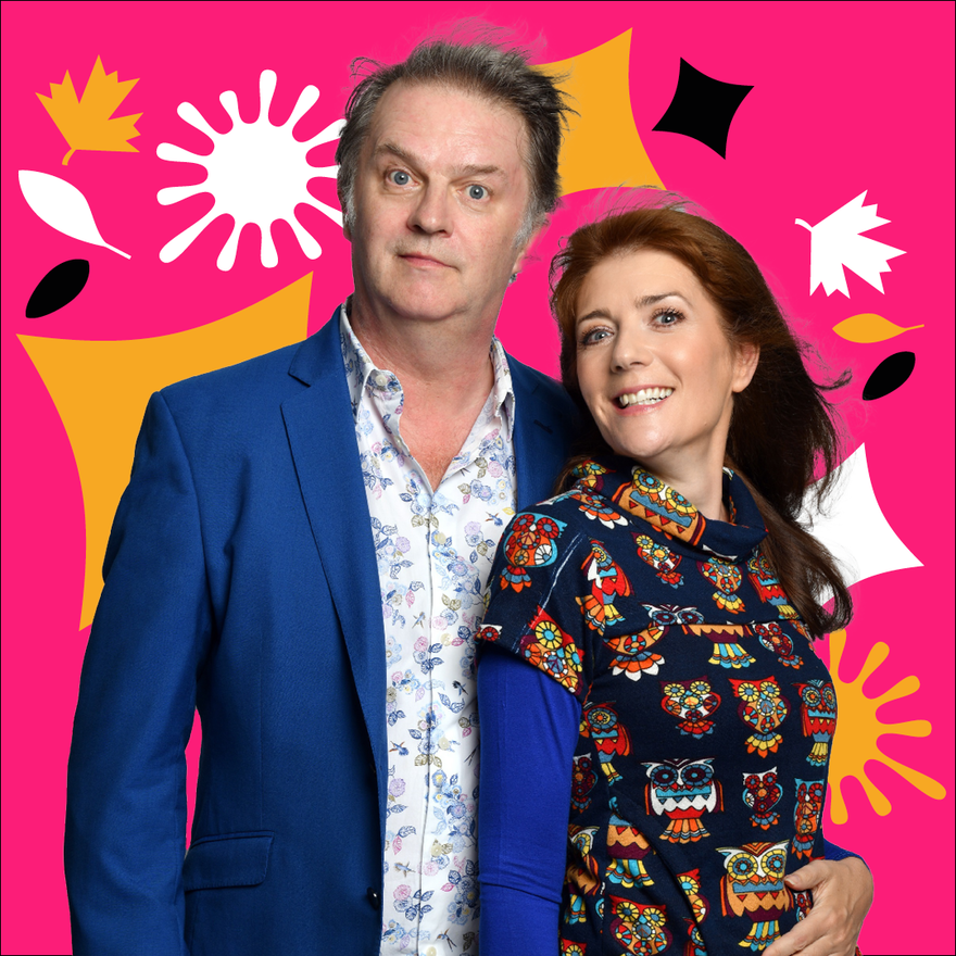 Paul Merton wearing a blue suit And Suki Webster a multi-coloured blouse standing in front of a pink background