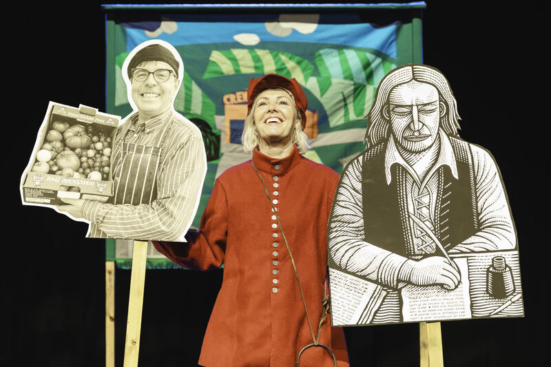 Victoria Melody on stage wearing a red medieval Royalist costume holding cut outs of Gerrard Winstanley and Brian, a Whitehawk based community food provider