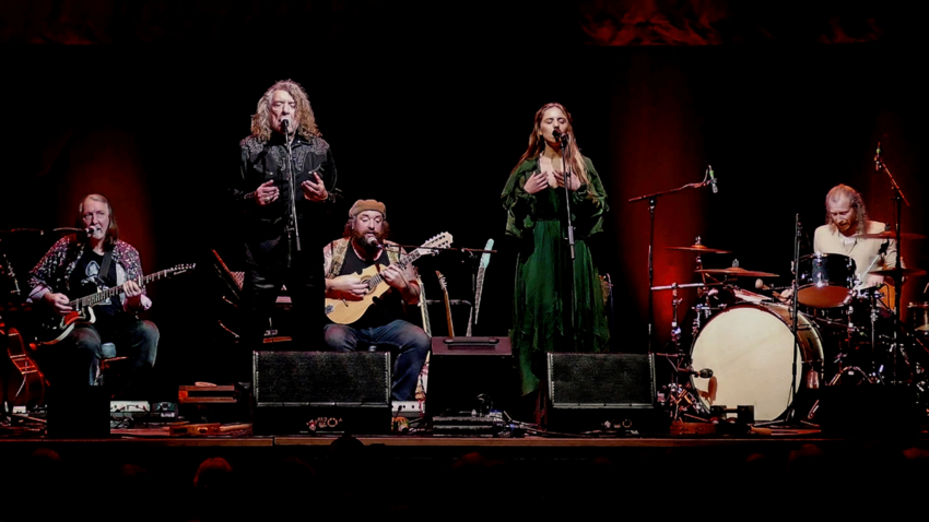 five people are on a stage performing. two sing and three in the back play their instruments