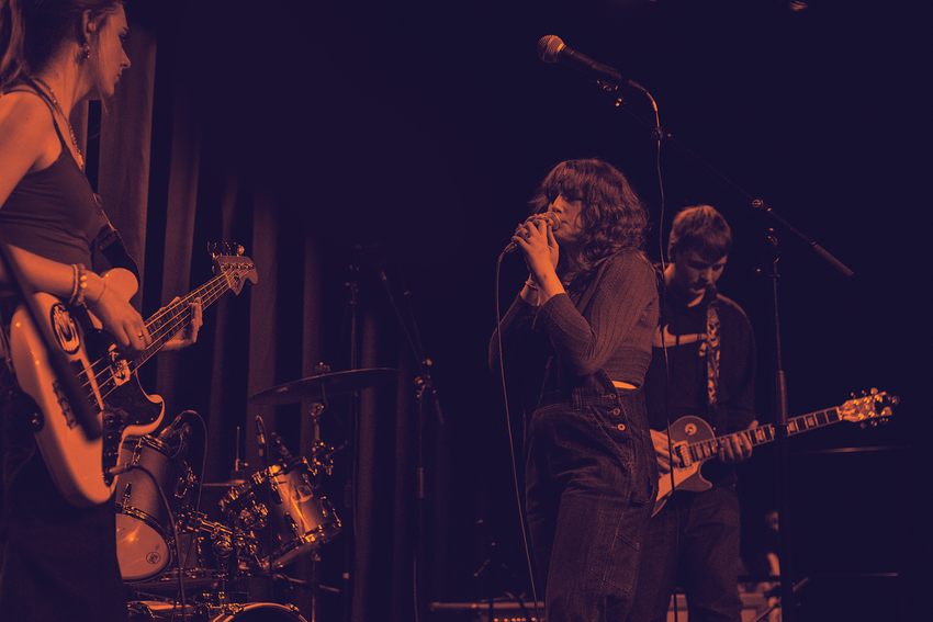 Teenage musicians performing on stage. One is playing guitar, one is playing bass, one is singing into a mic and one is playing the drums. The image has a colour treatment in the Create Music branding
