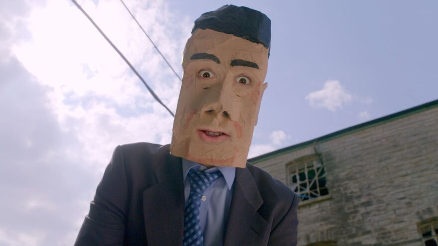 A person wearing a giant paper mache head looks down to the camera. Behind them is a bright blue sky and a white brick building 