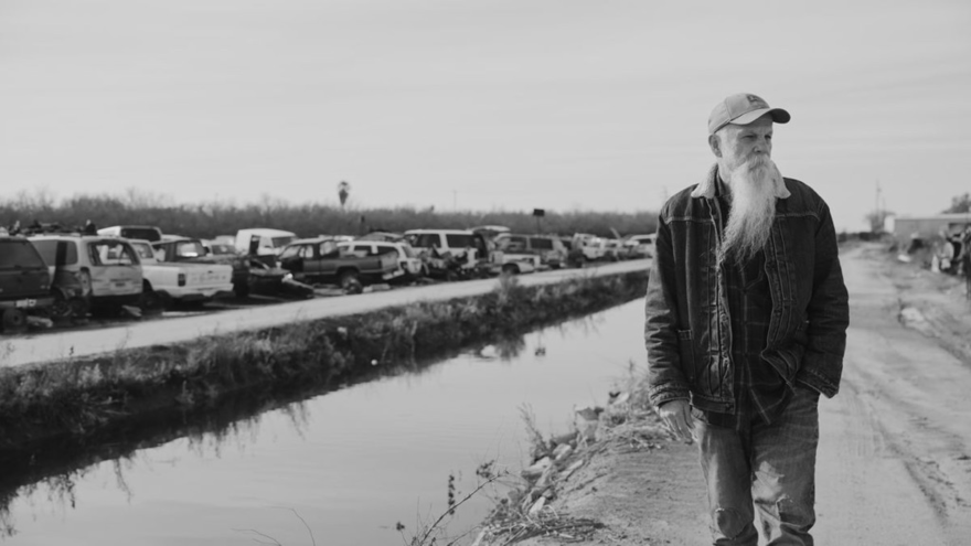 Black and White image of a man with a white beard walking a long a canal
