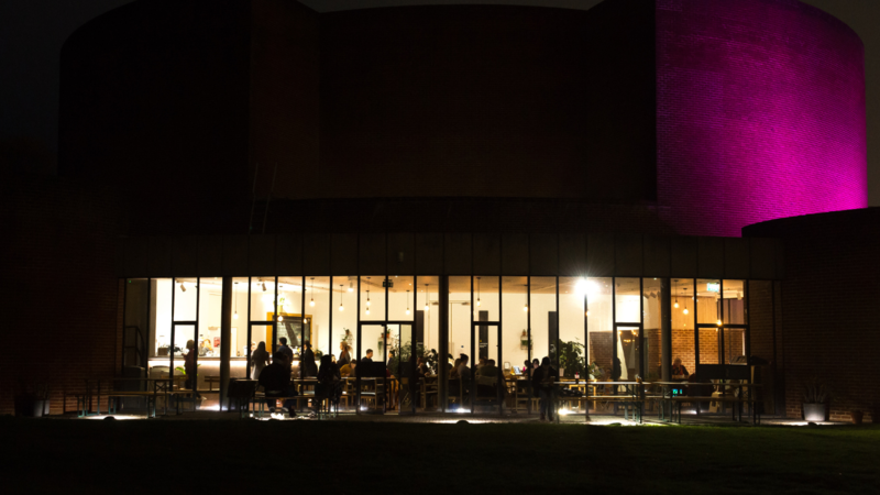 Exterior of a building at night, inside the building the lights are on, showing off large windows and a busy cafe space