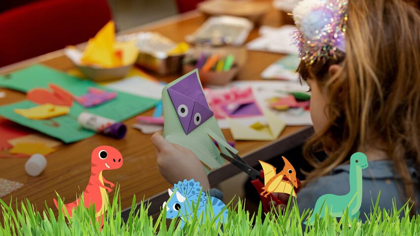 Close up image taken from behind a little girl's shoulder as she cuts paper. Cartoon dinosaurs and grass are layered over the top of the photo