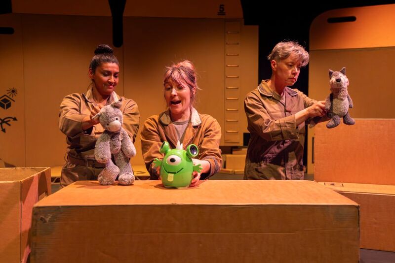 Three people holding toys up, they are smiling. They are surrounded by cardboard boxes