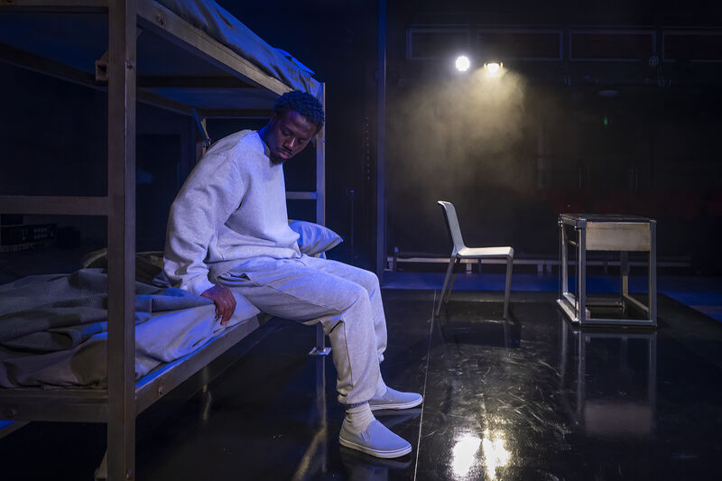An actor wearing a grey tracksuit sits on the bottom bunk of a metal bunk bed, he wears a sad expression on his face