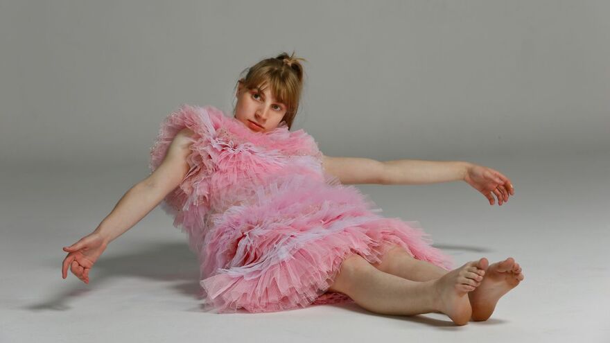 A woman in a bright pink tulle dress is sat on the floor and leaning backwards. She is in a plain white room
