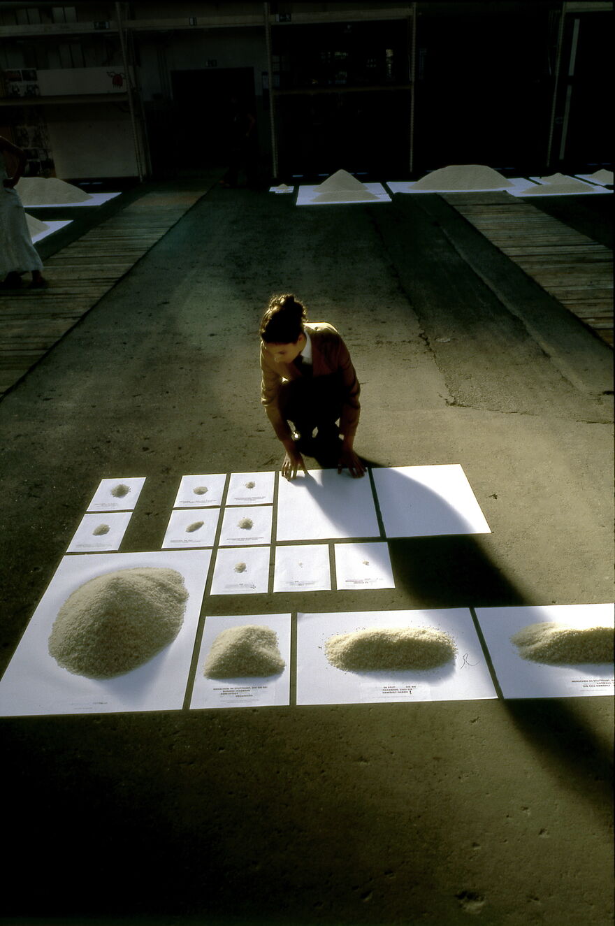 Sheets of white paper lie on the floor of a warehouse. On each sheet there's a different sized pile of rice with writing beneath. A performer leans over an empty sheet