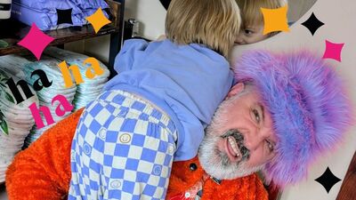 Rich Wilson wearing a purple fuzzy hat with an angry and confused facial expression. He is holding a young child over his shoulder. He is surrounded by black, yellow and pink stars and 'ha ha ha'