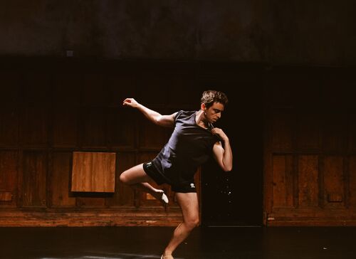 A person dancing on a dimly lit stage. One arm is up in the air and the other is touching his shoulder. He is standing on one bent leg and looks sad