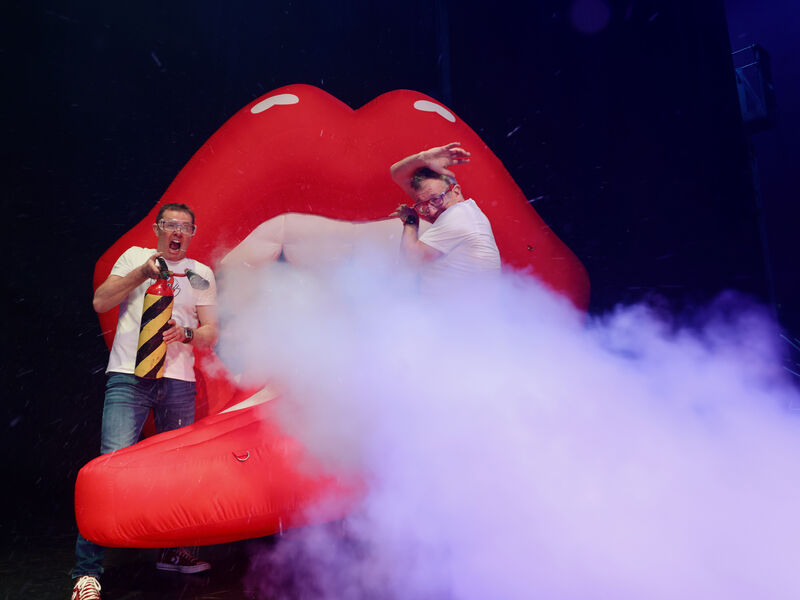 One man sprays a fire extinguisher in front of a giant inflatable mouth while the other man ducks away from the fog
