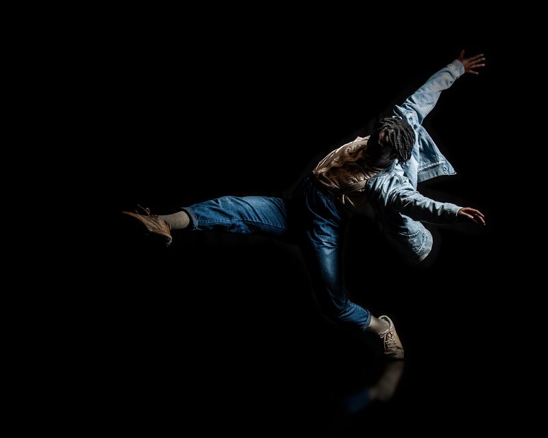 A dancer in jeans and denim jacket, with a leg elegantly kicked forwards and arms raised behind from the momentum of kicking forward