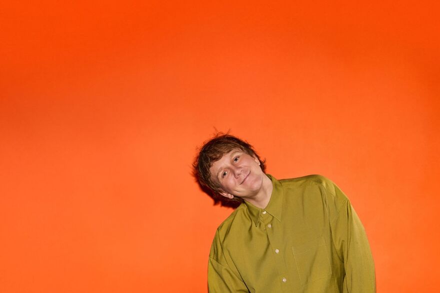Josh Thomas is wearing a msutard coloured shirt in front of a bright orange background, he is tilting to the left