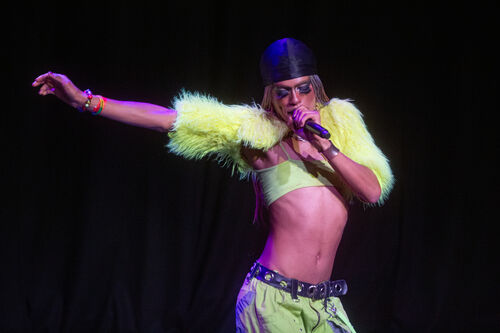 A performer against a black background. They are singing into a microphone and wearing a neon green fluffy jacket.