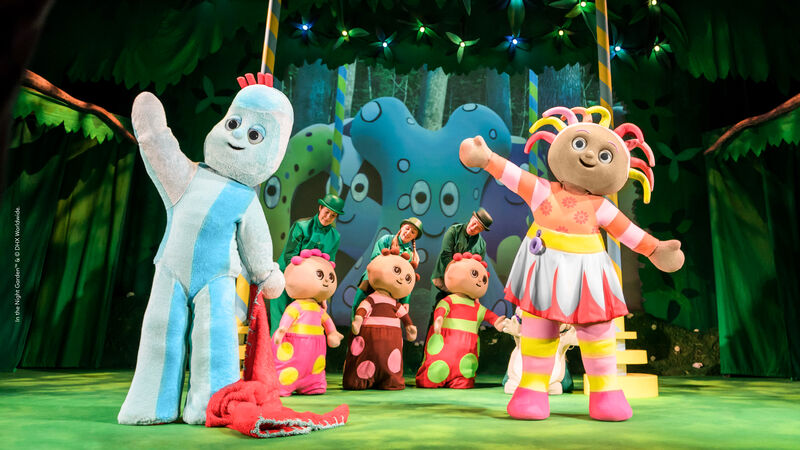 A large costumed blue creature and large costumed figure wearing a multi-coloured dress stand waving with puppeteers controlling three round figures in the centre background