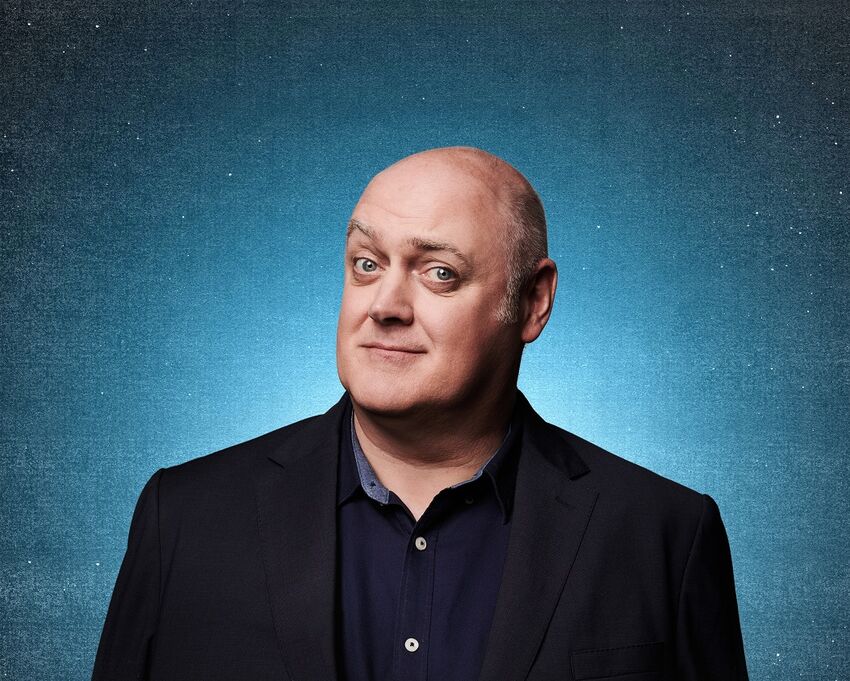 A white, bald man in a black suit jacket and black shirt with an open collar