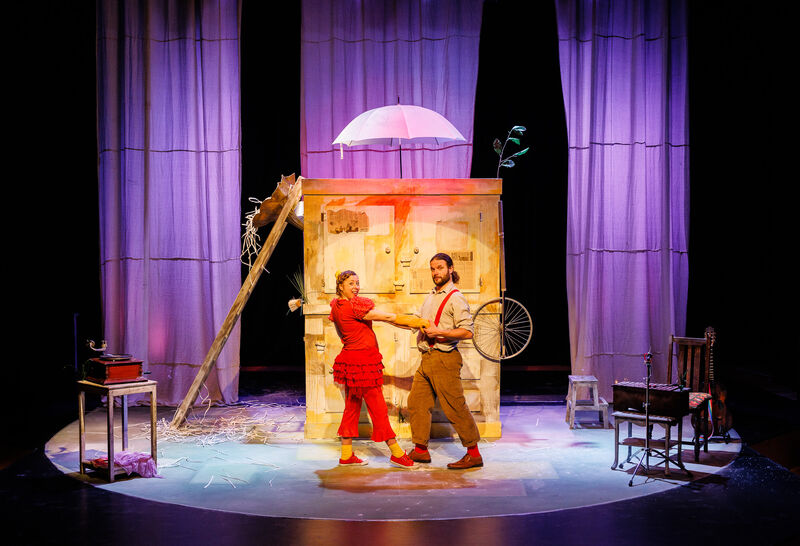 A woman dressed as a red hen and a man face each other on stage in front of a tall wooden box