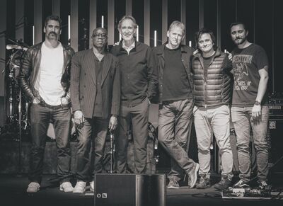 Group of 6 men wearing jeans and various t-shirts, gilets and jackets. They are standing on a stage and staring at the camera. The photo is black and white
