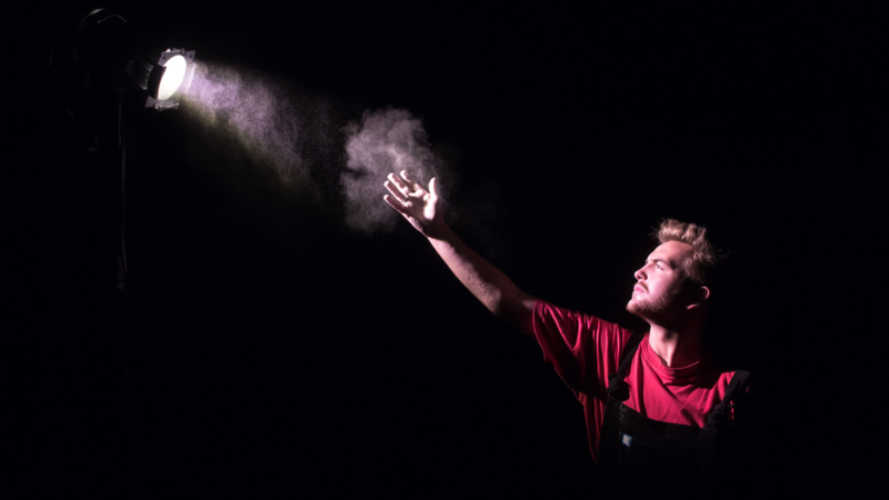 A white man with blonde hair is lit by a white spotlight. He wears a red long-sleeved t-shirt. His right arm is raised up, with his open-palmed hand lit by the spotlight,
