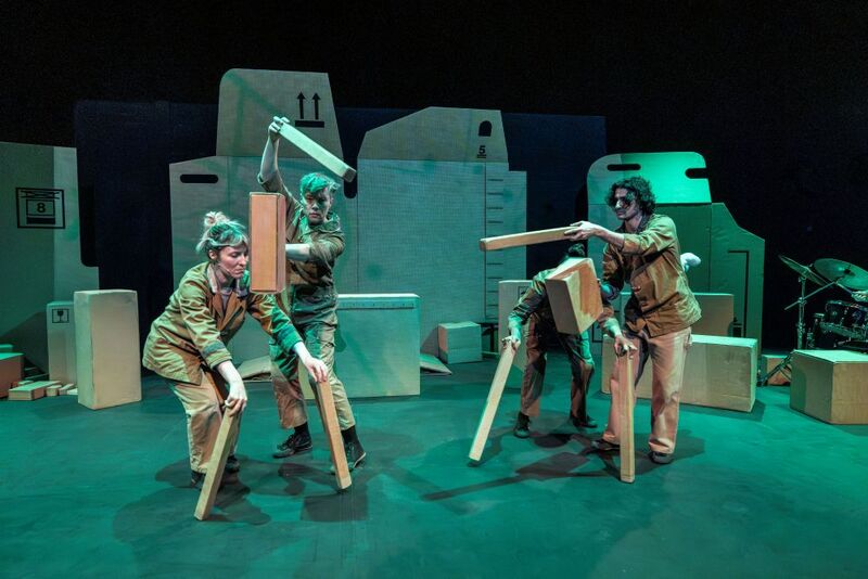 Four people on stage holding different cardboard shapes. They are on stage doing a performance surrounded by cardboard boxes