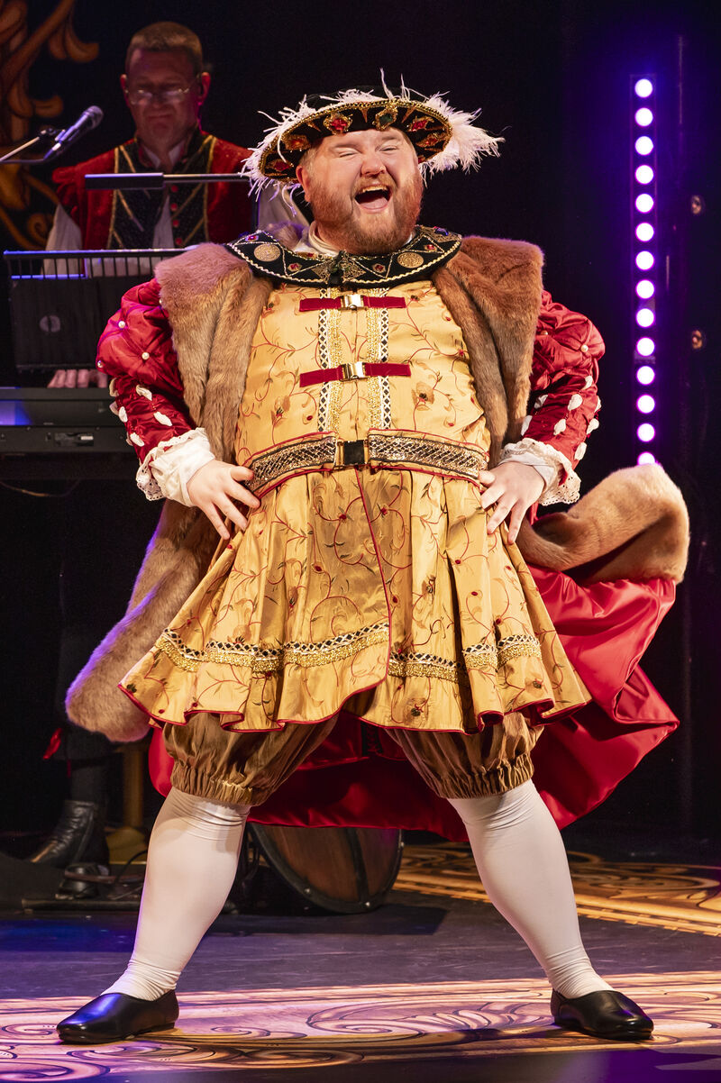 A man dressed as Henry 8th stands with legs shoulder width apart with his hands on his hips
