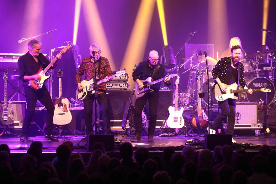 Four men on stage playing guitars. The stage is lit with yellow and purple stage lights